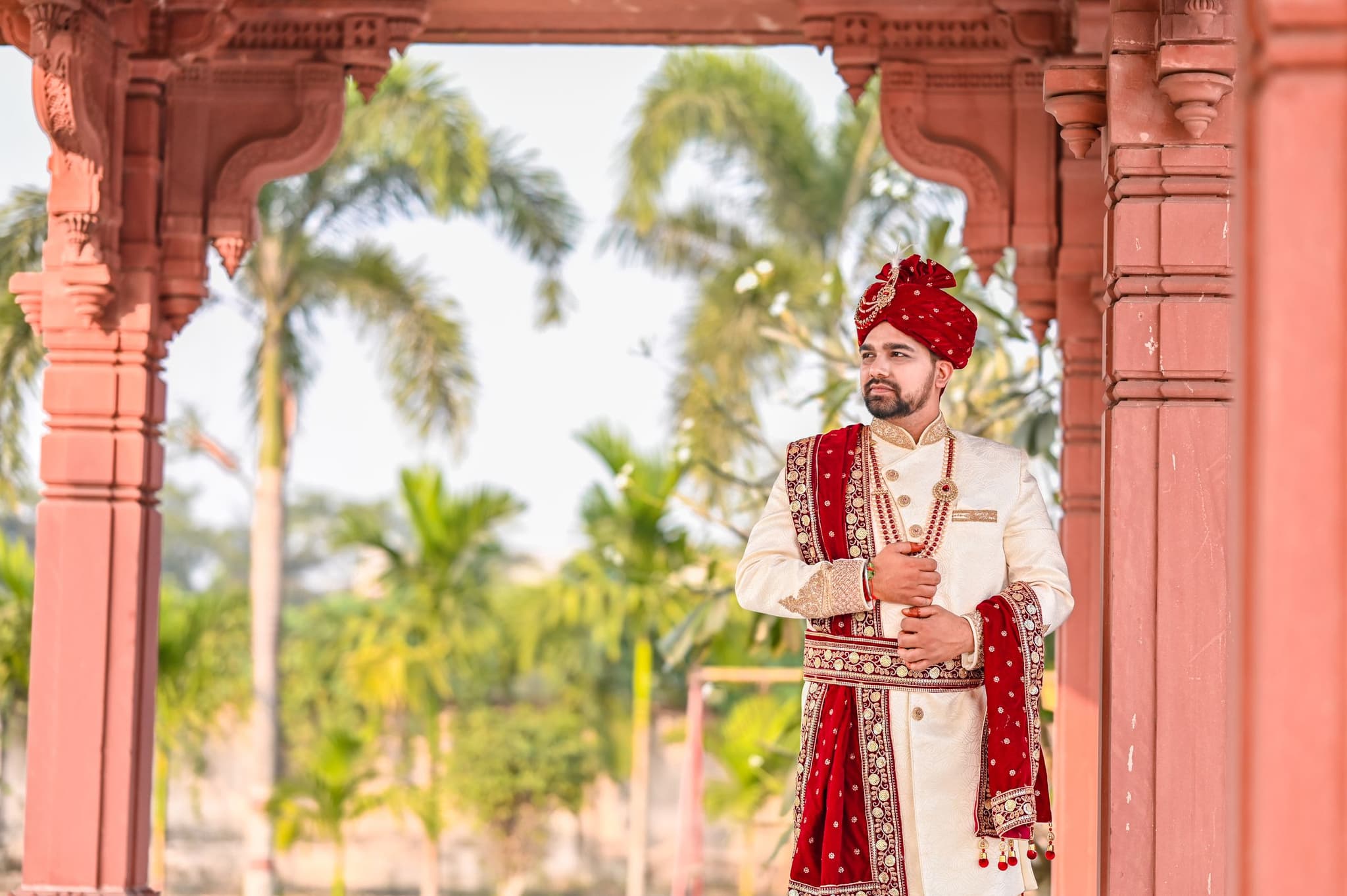 A groom in an ivory sherwani, red turban and embroidered red shawl beneath a carved archway